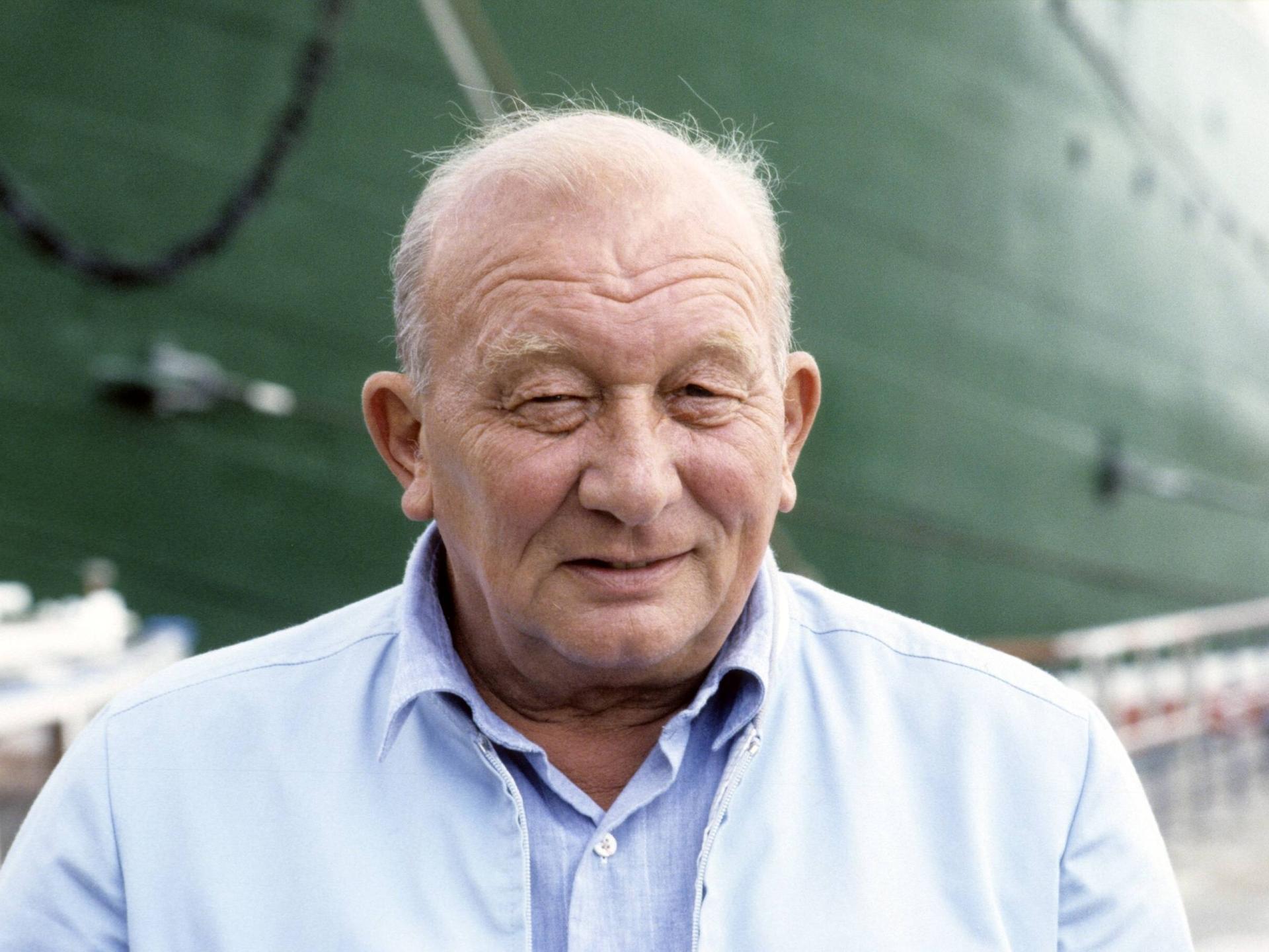 Der Schriftsteller James Krüss 1988. Er steht vor einem Schiff mit dunkelgrünem Anstrich und lächelt verschmitzt. 