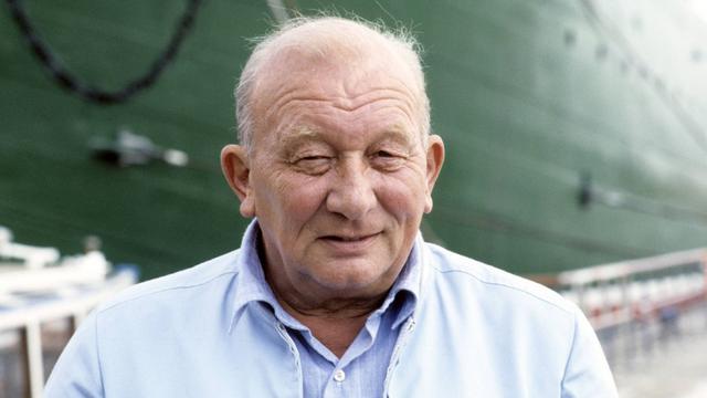 Der Schriftsteller James Krüss 1988. Er steht vor einem Schiff mit dunkelgrünem Anstrich und lächelt verschmitzt. 