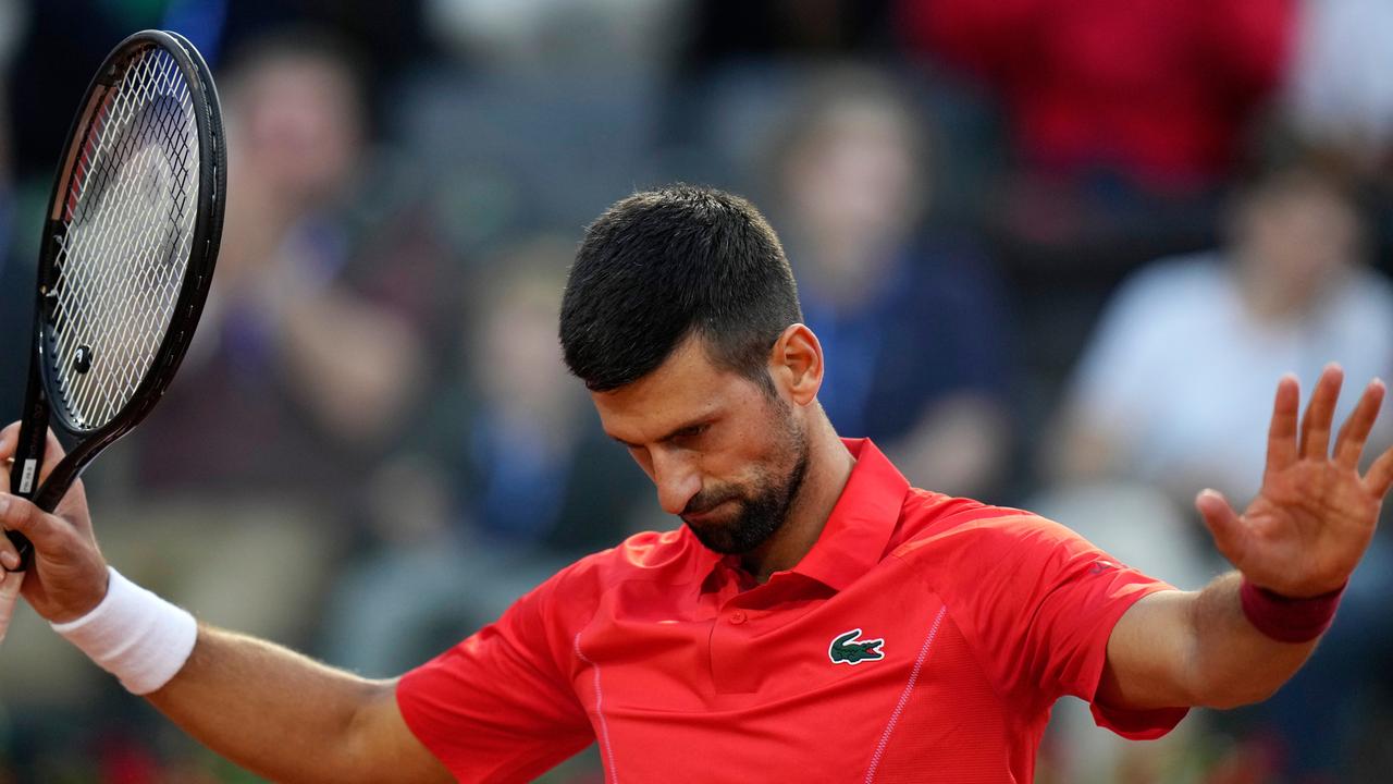 Der Tennisspieler Novak Djokovic hebt beide Arme zur Seite. In der rechten Hand hält er den Tennisschläger. Sein Kopf ist ein bisschen gesenkt. Djokovic trägt ein rotes Trikot. 