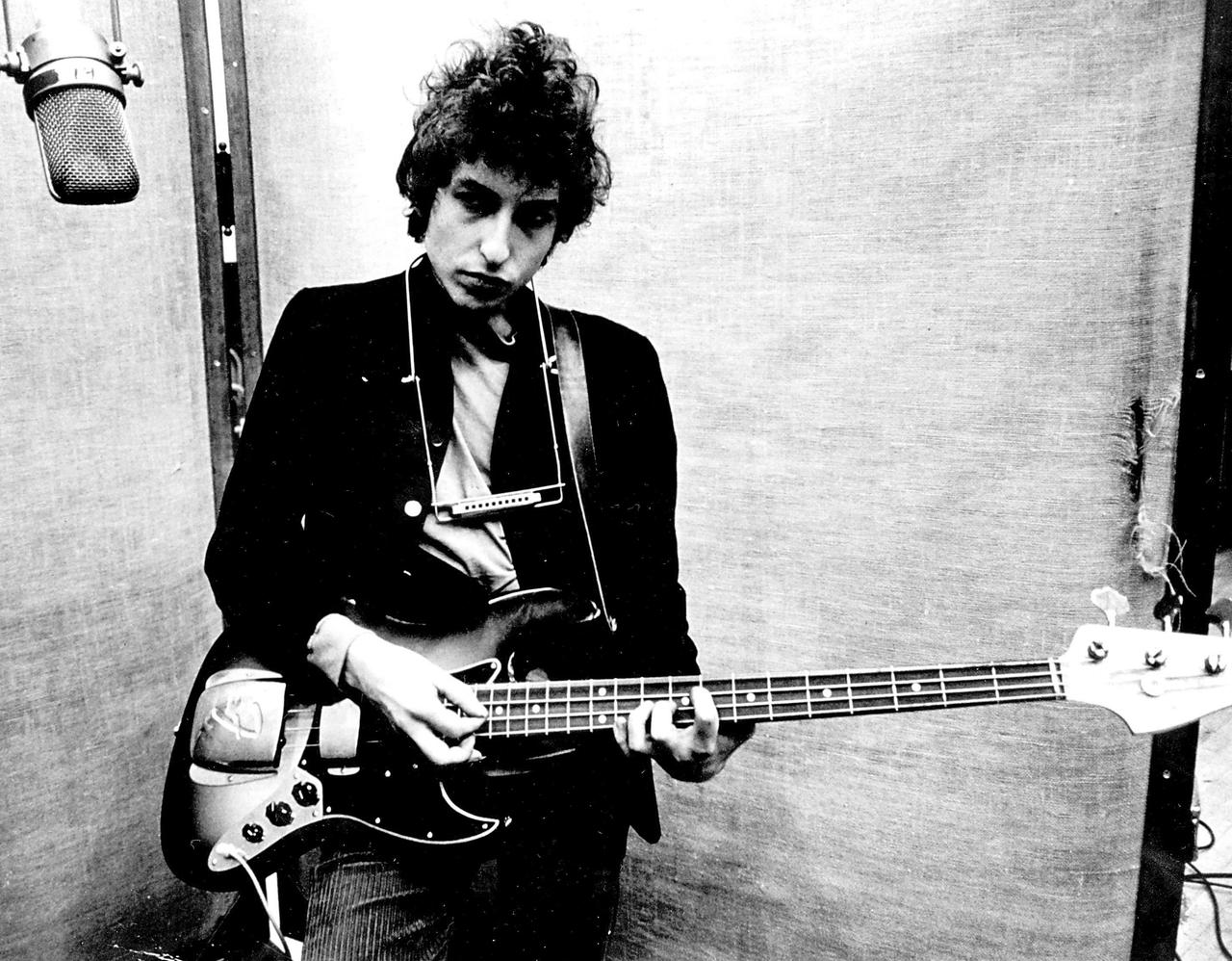 Die Schwarzweißaufnahme zeigt Bob Dylan 1965 in einem Tonstudio in New York mit einem E-Bass und mit Mundharmonika um den Hals.