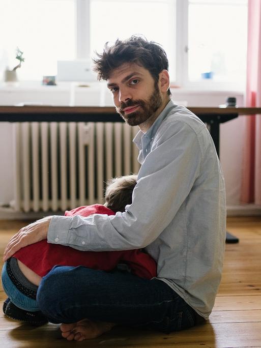 Der Schriftsteller Tobias Wilhelm sitzt mit verschränkten Beinen auf dem Boden, mit abgewandtem Gesicht liegt ein Kind in seinem Schoß, im Hintergrund sind ein Schreibtisch und Gardinen zu sehen. 