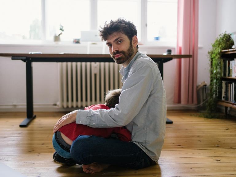 Der Schriftsteller Tobias Wilhelm sitzt mit verschränkten Beinen auf dem Boden, mit abgewandtem Gesicht liegt ein Kind in seinem Schoß, im Hintergrund sind ein Schreibtisch und Gardinen zu sehen. 