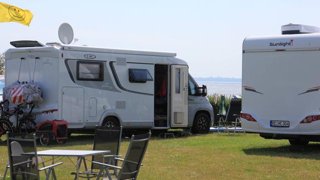Zwei Wohn-Mobile stehen nebeneinander auf dem Rasen. Im Hinter-Grund ist die Ost-See zu sehen. Im Vorder-Grund stehen ein Camping-Tisch und Camping-Stühle. 