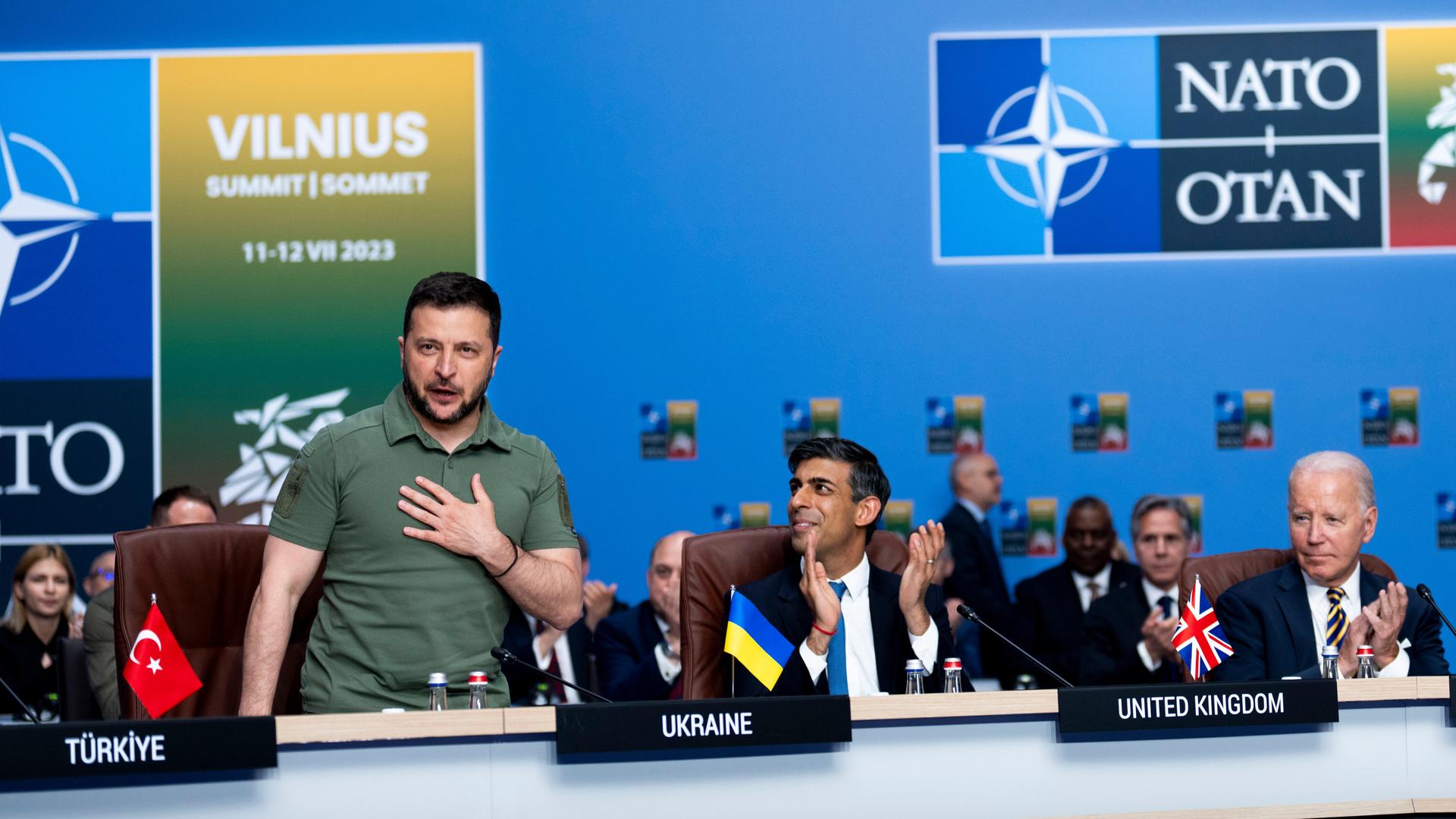 Ukraines Präsident Wolodymyr Selenskyj(l.) steht beim NATO-Gipfel im litauischen Vilnius an seinem Platz und bekommt Applaus von NATO-Mitgliedern, darunter Großbritanniens Premierminister Rishi Sunak und US-Präsident Joe Biden (r.)