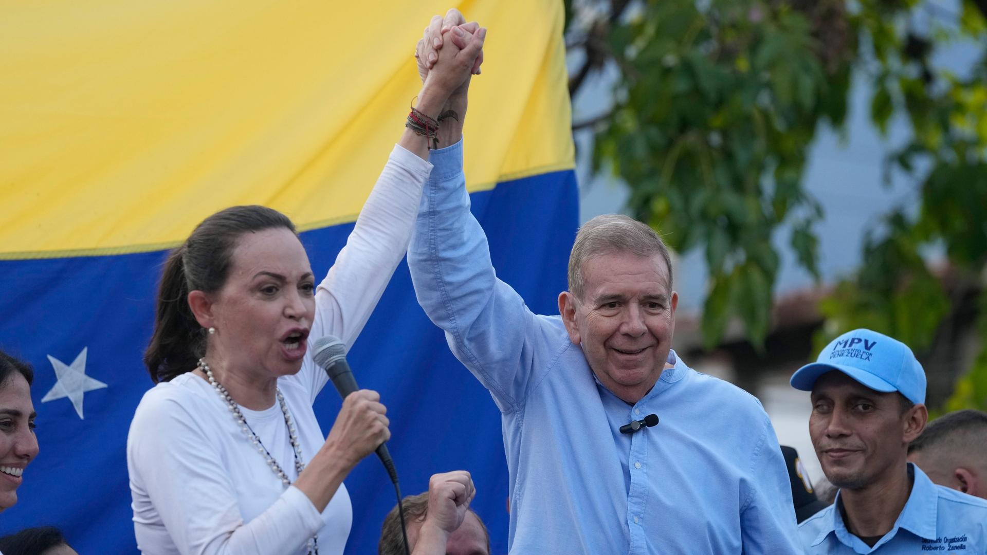 Maria Corina Machado und Edmundo Gonzalez Urrutia recken ihre Hände nach oben. Machado hält ein Mikrofon in die Höhe, im Hintergrund die venezolanische Flagge.