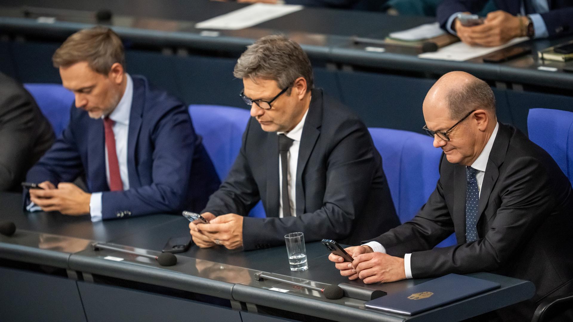 Christian Lindner, Robert Habeck und Olaf Scholz sitzen nebeneinander im Bundestag. Jeder von ihnen blickt ins eigene Handy.
