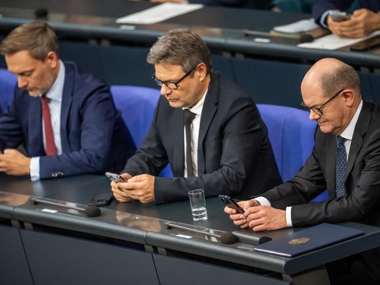 Christian Lindner, Robert Habeck und Olaf Scholz sitzen nebeneinander im Bundestag. Jeder von ihnen blickt ins eigene Handy.