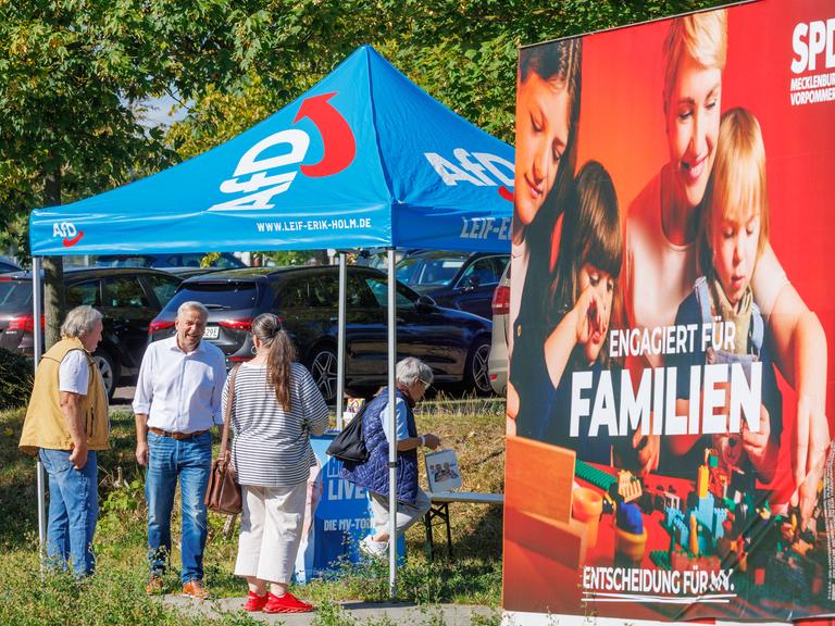 Leif-Erik Holm, AfD-Landesvorsitzender in Mecklenburg-Vorpommern, spricht an einem Informationsstand der Partei mit Passanten. Im Vordergrund ist ein Wahlplakat der SPD zu sehen. 