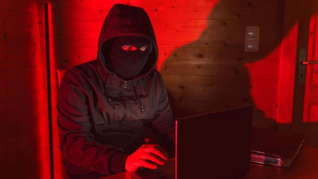 Ein maskierter Mann sitz am Laptop. Neben ihm liegt ein Smarthpone.