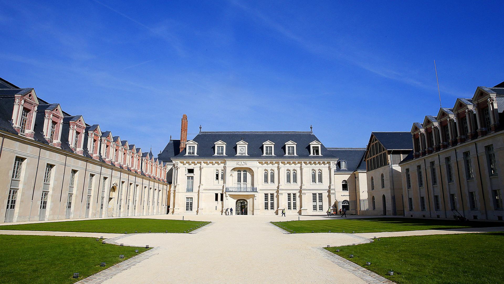 Die Frontansicht auf das Schloss von Villers-Cotterêts, einem historischen Gebäude mit heller Fassade und zwei Flügeln, vor dem Eingang befindet sich breite Schotterwege und Wiesen.