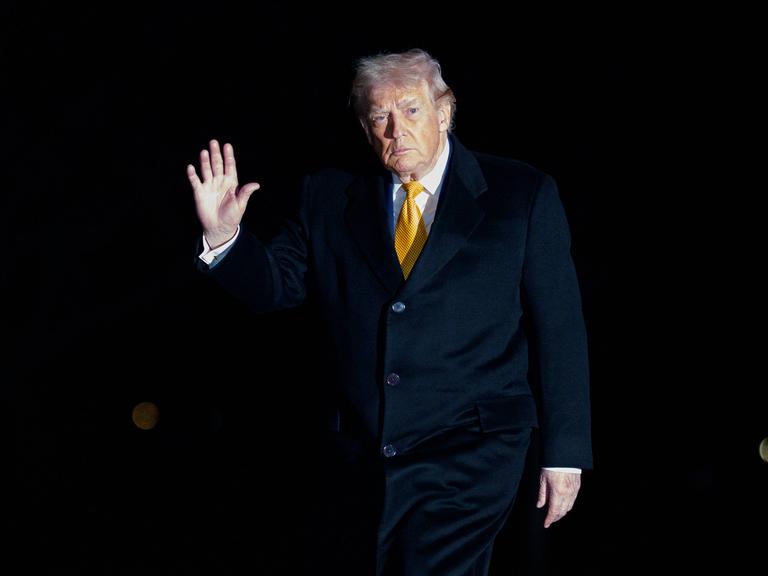 Donald Trump steht im Dunkeln, nur sein Gesicht und die zum Gruß erhobene Hand sind zu sehen