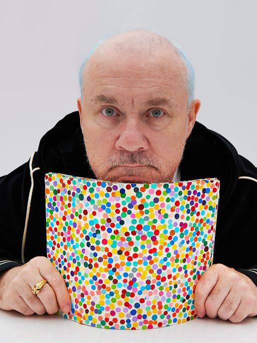 Der Künstler Damien Hirst, in Trainingsjacke und mit einem goldenen Ring an der rechten Hand, präsentiert ein bunt gepunktetes Bild aus seiner NFT-Serie "The Currency".