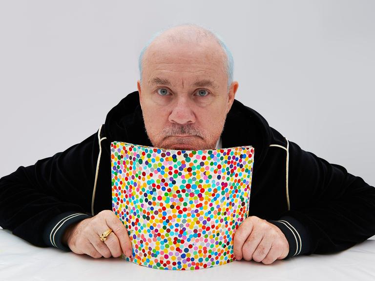 Der Künstler Damien Hirst, in Trainingsjacke und mit einem goldenen Ring an der rechten Hand, präsentiert ein bunt gepunktetes Bild aus seiner NFT-Serie "The Currency".