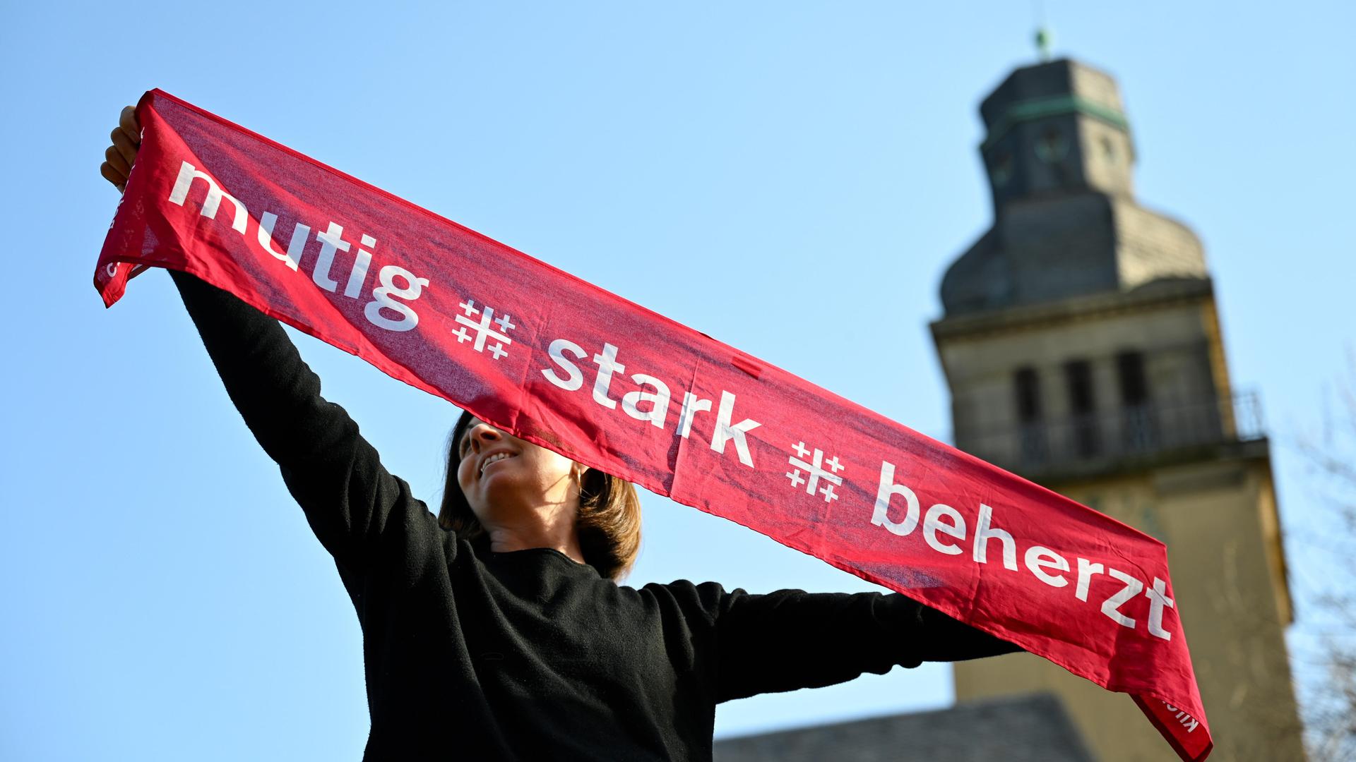 Eine junge Frau hält einen Kirchentagsschal mit der Losung "mutig - stark - beherzt" zum Deutschen Evangelischen Kirchentag über ihren Kopf, im Hintergrund rechts sieht man einen Kirchturm. Eine junge Frau hält einen Kirchentagsschal mit der Losung "mutig - stark - beherzt" zum Deutschen Evangelischen Kirchentag über ihren Kopf, im Hintergrund rechts sieht man einen Kirchturm.