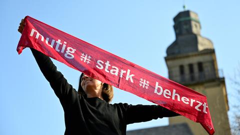 Eine junge Frau hält einen Kirchentagsschal mit der Losung "mutig - stark - beherzt" zum Deutschen Evangelischen Kirchentag über ihren Kopf, im Hintergrund rechts sieht man einen Kirchturm.