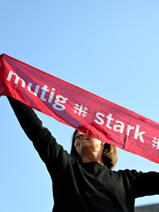 Eine junge Frau hält einen Kirchentagsschal mit der Losung "mutig - stark - beherzt" zum Deutschen Evangelischen Kirchentag über ihren Kopf, im Hintergrund rechts sieht man einen Kirchturm.