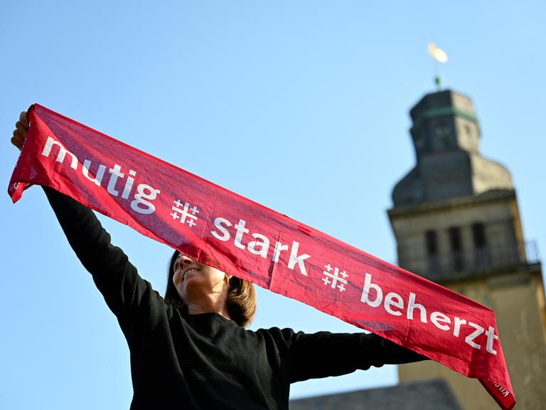 Eine junge Frau hält einen Kirchentagsschal mit der Losung "mutig - stark - beherzt" zum Deutschen Evangelischen Kirchentag über ihren Kopf, im Hintergrund rechts sieht man einen Kirchturm.