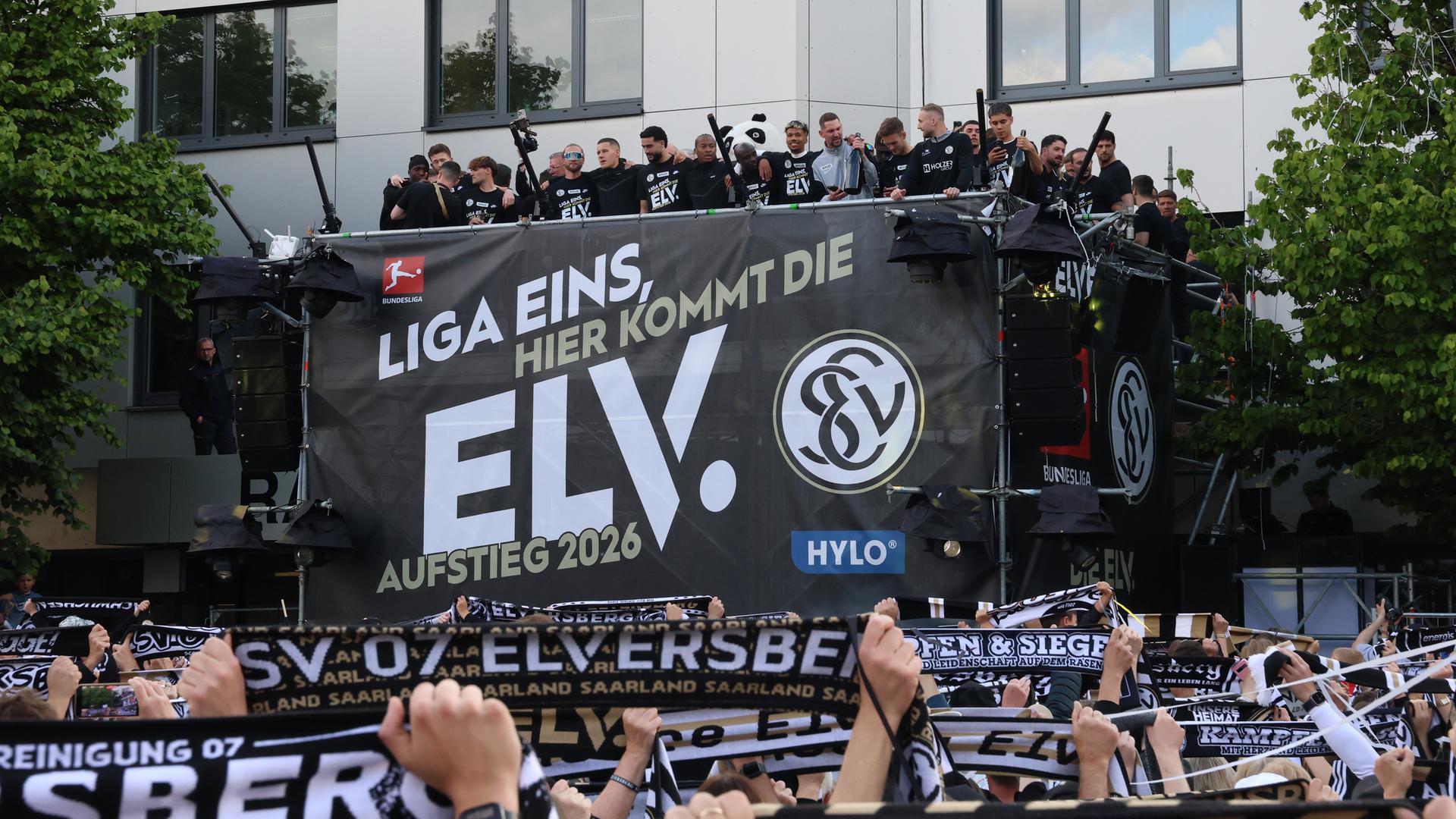 Auf einer Bühne steht die Mannschaft der SV Elversberg. Im Vordergrund halten Fans ihre Hände in die Höhe.