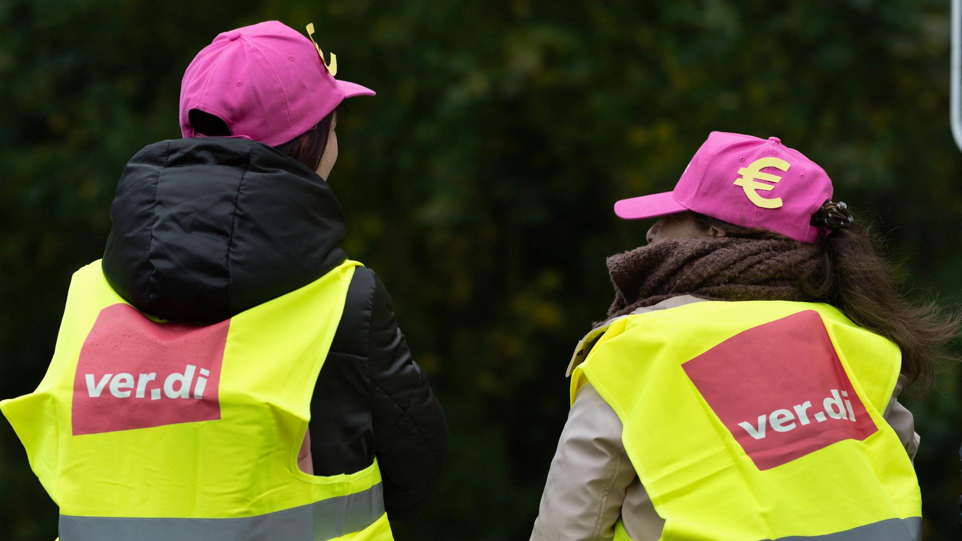 Zwei Frauen sitzen mit dem Rücken zum Betrachter. Sie tragen neongelbe Westen mit dem Verdi-Logo und magentafarbene Kappen mit einem Euro-Zeichen.