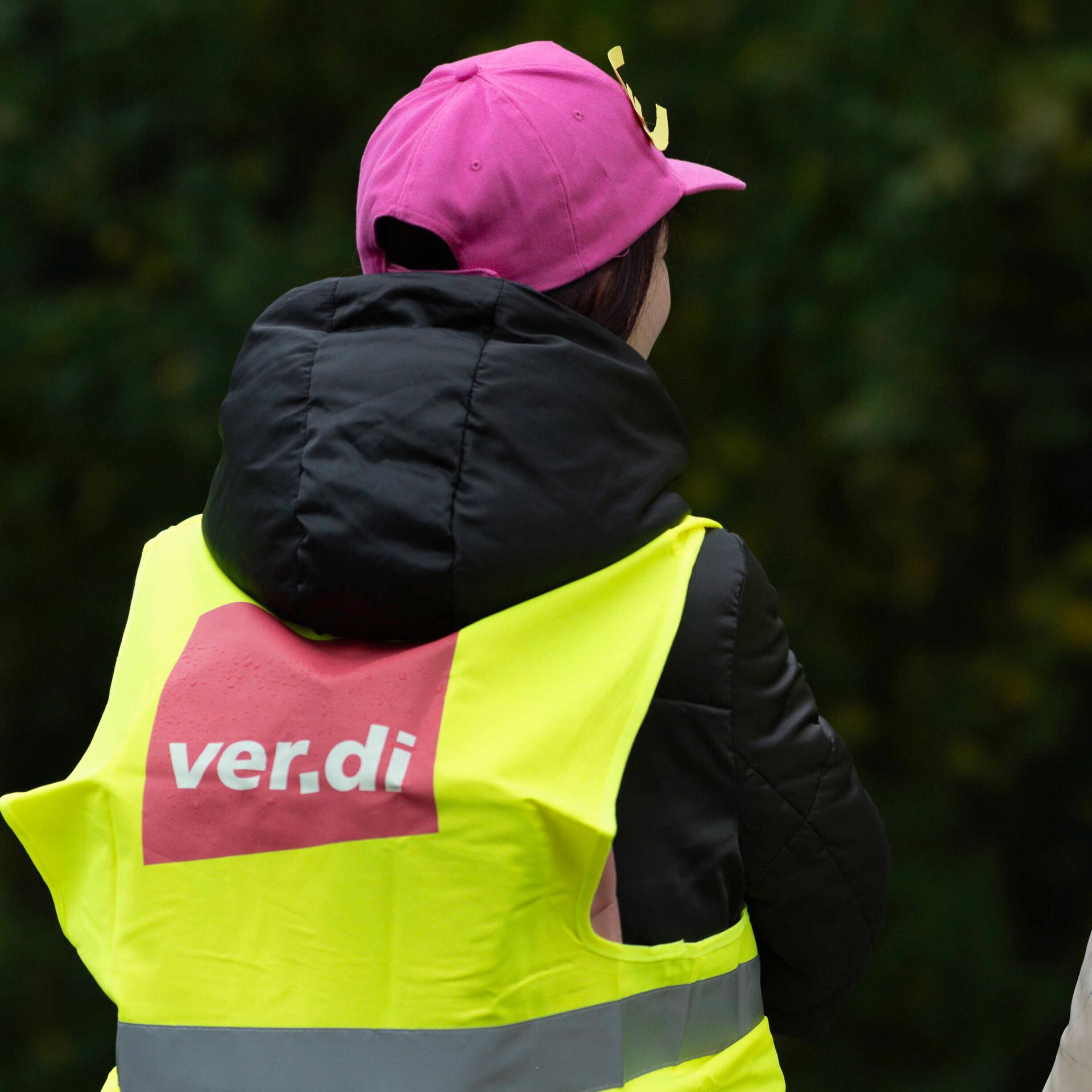 Zwei Frauen sitzen mit dem Rücken zum Betrachter. Sie tragen neongelbe Westen mit dem Verdi-Logo und magentafarbene Kappen mit einem Euro-Zeichen.