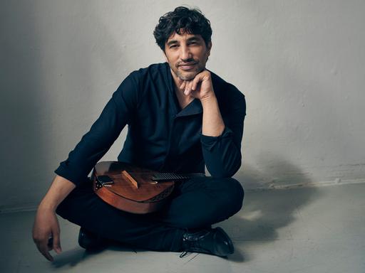Avi Avital sitzt im Schneidersitz vor der Kamera und lächelt, während seine Mandoline im Schoß liegt.