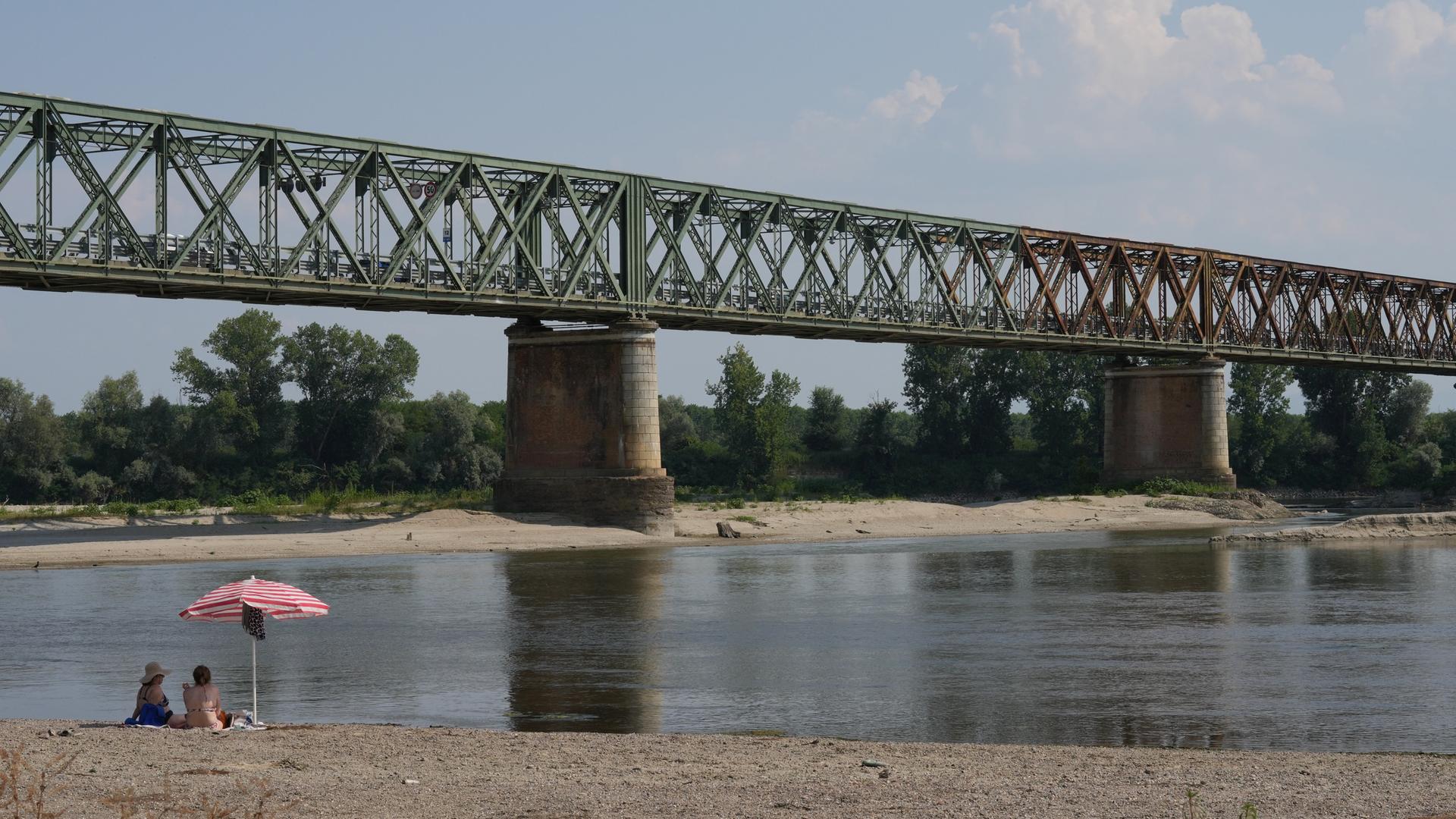 Blick auf ein Stück des Flusses unter einer Eisenbahnbrücke mit relativ wenig Wasser. Am vorderen Bildrand sitzen zwei Menschen unter einem Sonnenschirm. Blick auf ein Stück des Flusses unter einer Eisenbahnbrücke mit relativ wenig Wasser. Am vorderen Bildrand sitzen zwei Menschen unter einem Sonnenschirm.