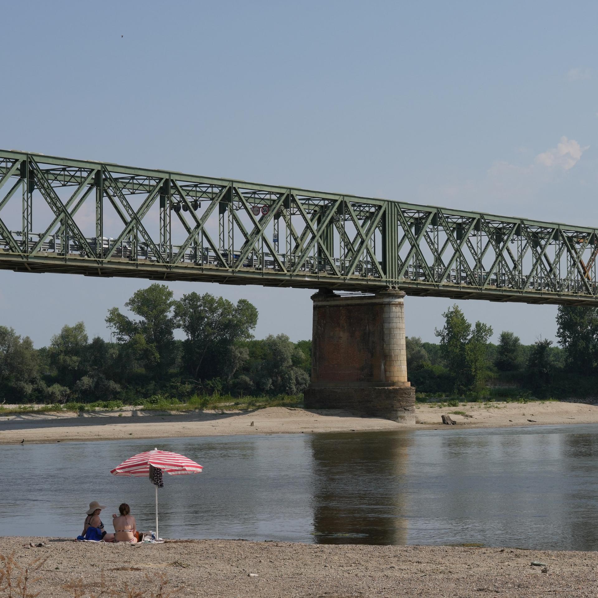 Blick auf ein Stück des Flusses unter einer Eisenbahnbrücke mit relativ wenig Wasser. Am vorderen Bildrand sitzen zwei Menschen unter einem Sonnenschirm. 