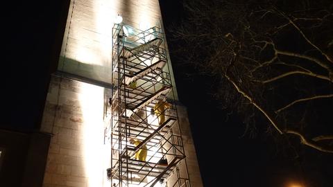 Zwei Männer in gelben Anzügen auf einem Gerüst am nächtlichen Luther Turm in Köln