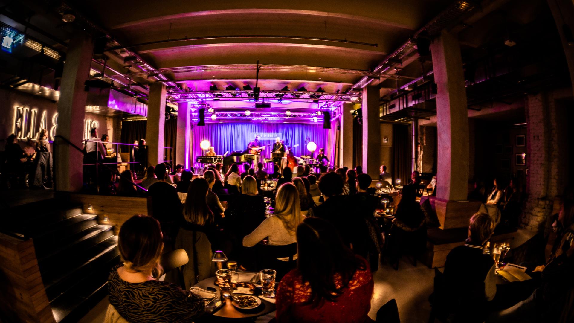 Blick in den Mannheimer Jazzclub "Ella & Louis", in dem viele Menschen an kleinen Tischen sitzen mit Getränken und auf die lile erleuchtete Bühne schauen.