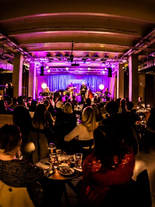 Blick in den Mannheimer Jazzclub "Ella & Louis", in dem viele Menschen an kleinen Tischen sitzen mit Getränken und auf die lile erleuchtete Bühne schauen.