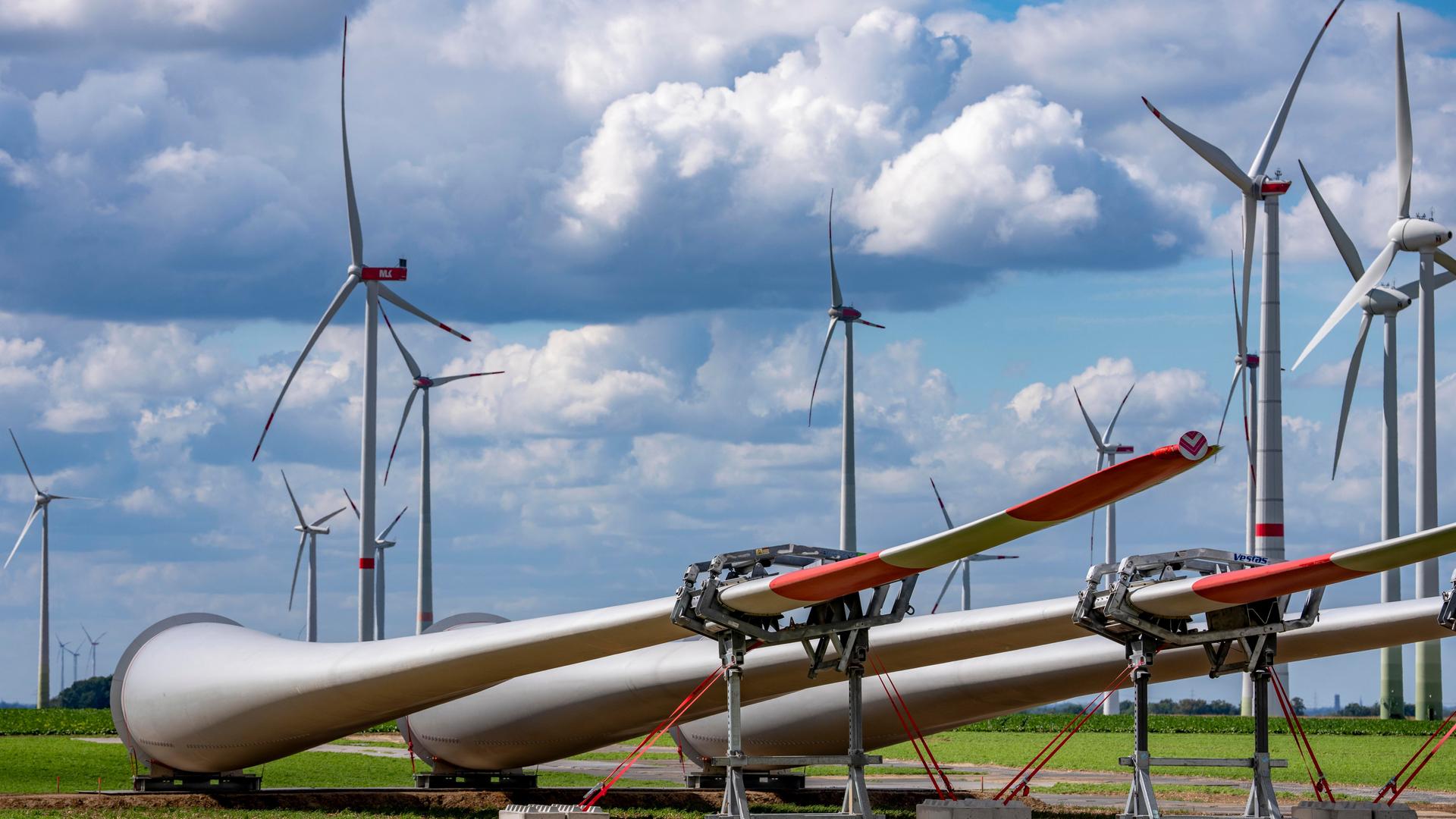 Mehrere große Windkraftanlagen stehen auf einem Feld. Im Vordergrund liegen Rotorblätter auf dem Boden, die für den Bau neuer Windräder vorbereitet werden. Über den Anlagen ziehen dichte Wolken am Himmel.