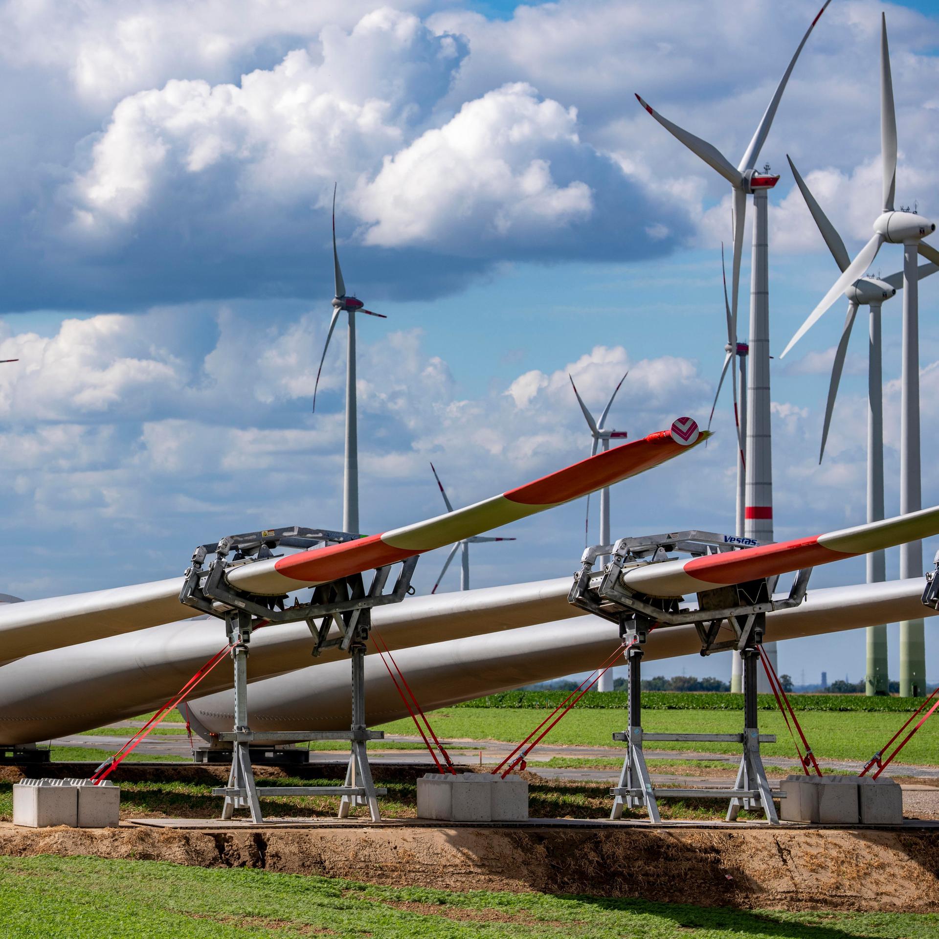 Mehrere große Windkraftanlagen stehen auf einem Feld. Im Vordergrund liegen Rotorblätter auf dem Boden, die für den Bau neuer Windräder vorbereitet werden. Über den Anlagen ziehen dichte Wolken am Himmel.