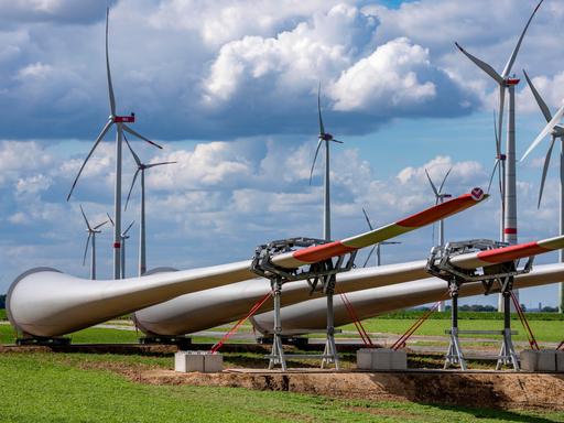 Mehrere große Windkraftanlagen stehen auf einem Feld. Im Vordergrund liegen Rotorblätter auf dem Boden, die für den Bau neuer Windräder vorbereitet werden. Über den Anlagen ziehen dichte Wolken am Himmel.