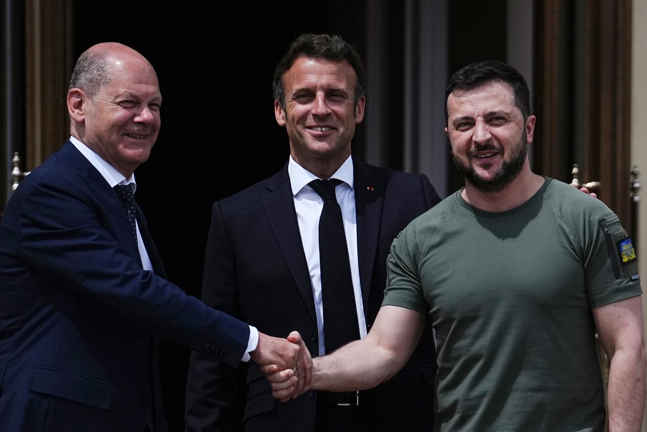 Bundeskanzler Olaf Scholz und der ukrainische Präsident Wolodymyr Selenskyj schütteln sich die Hand. In der Mitte steht der franzöische Präsident Emmanuel Macron.