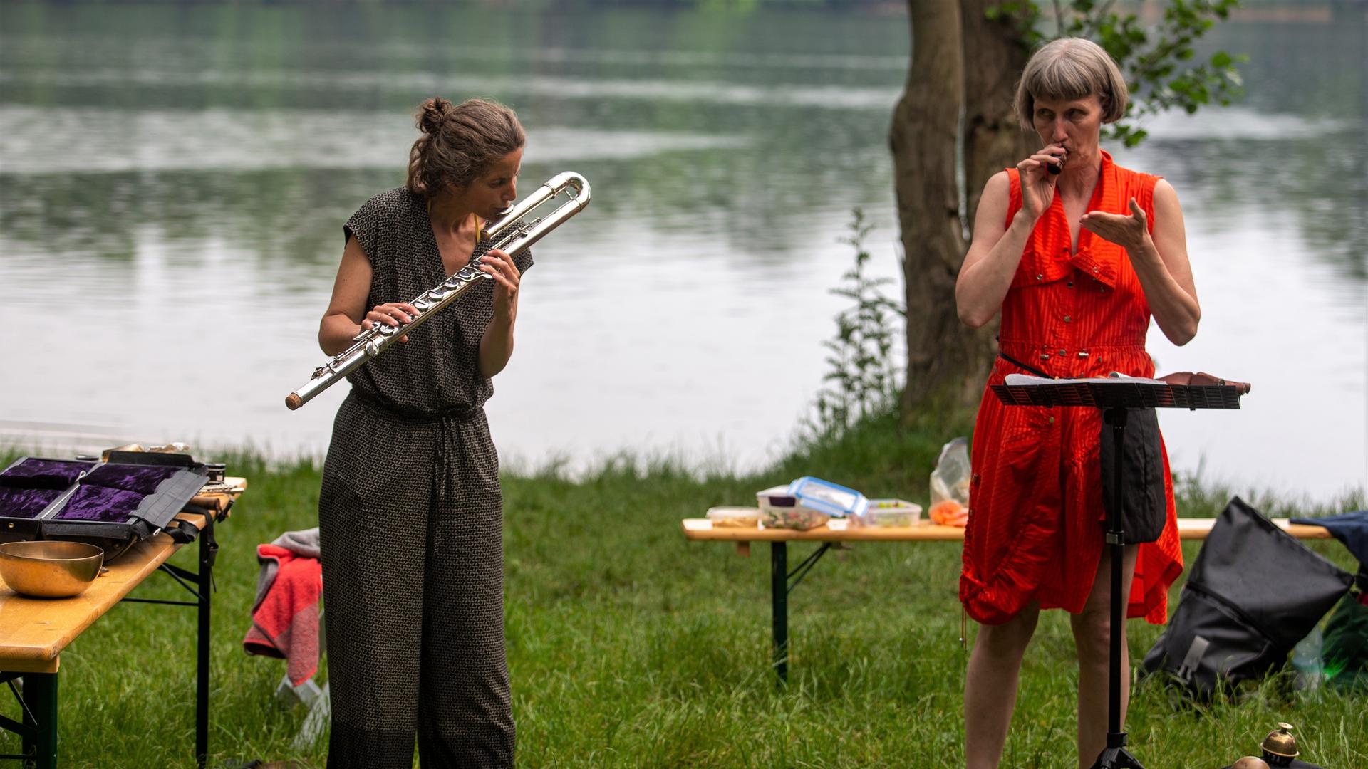 Sabine Vogel und Ute Wassermann stehen inmitten verschiedener Utensilien auf dem Rasen vor dem See und geben ein Konzert