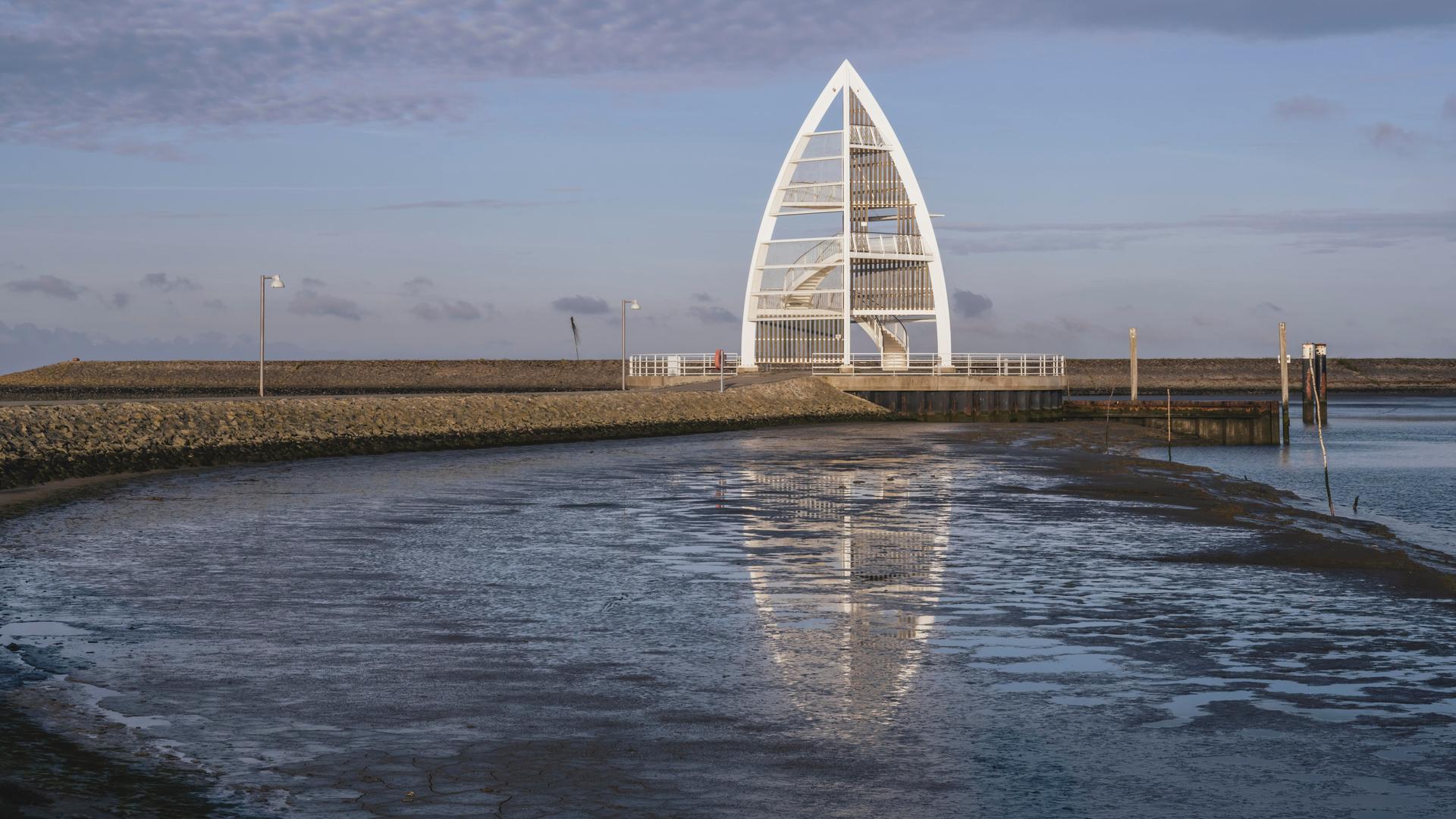 Ein Beobachtungsturm auf der Nordsee-Insel Juist. Im Turm führen sichtbar Treppen nach oben. Der Turm liegt auf einer Mole. Davor ist das Meer zu sehen. 