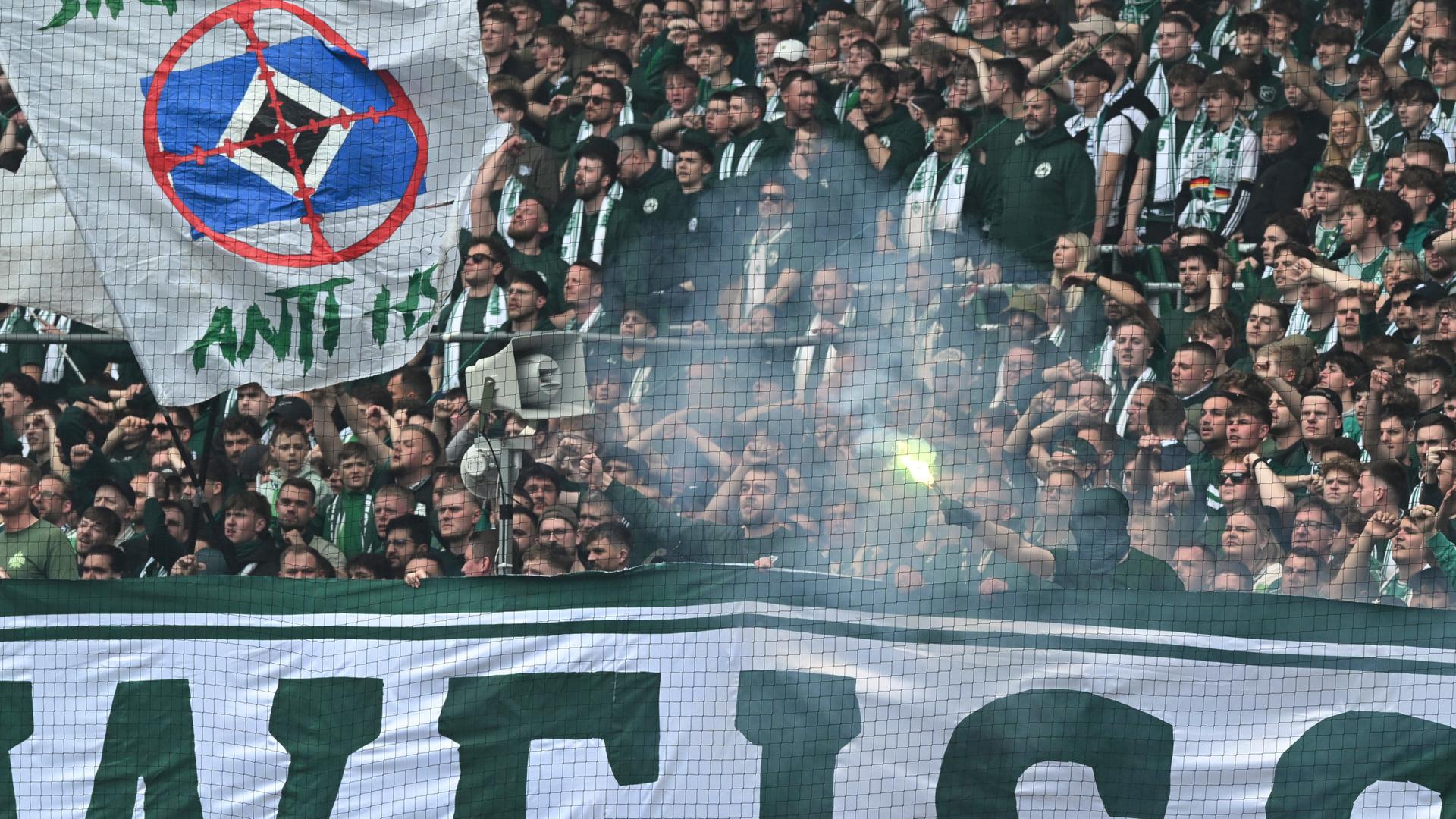 Werder Fans in der Ostkurve halten Fahnen und zünden pyrotechnische Gegenstände, aus denen Rauch aufsteigt. 