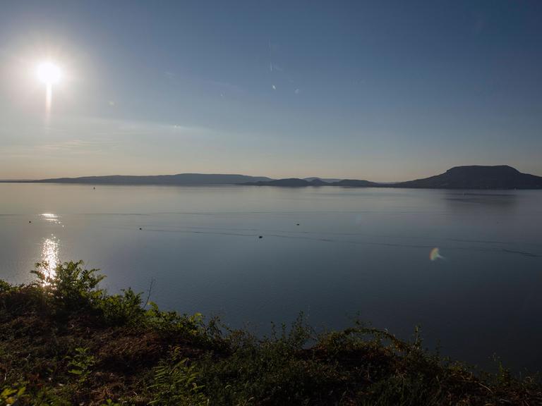 Das Bild blickt von der südlichen Seite auf den Plattensee. Der Himmel ist blau, anhand des Sonnenstandes kann man vom späten Nachmittag im Sommer ausgehen. 
