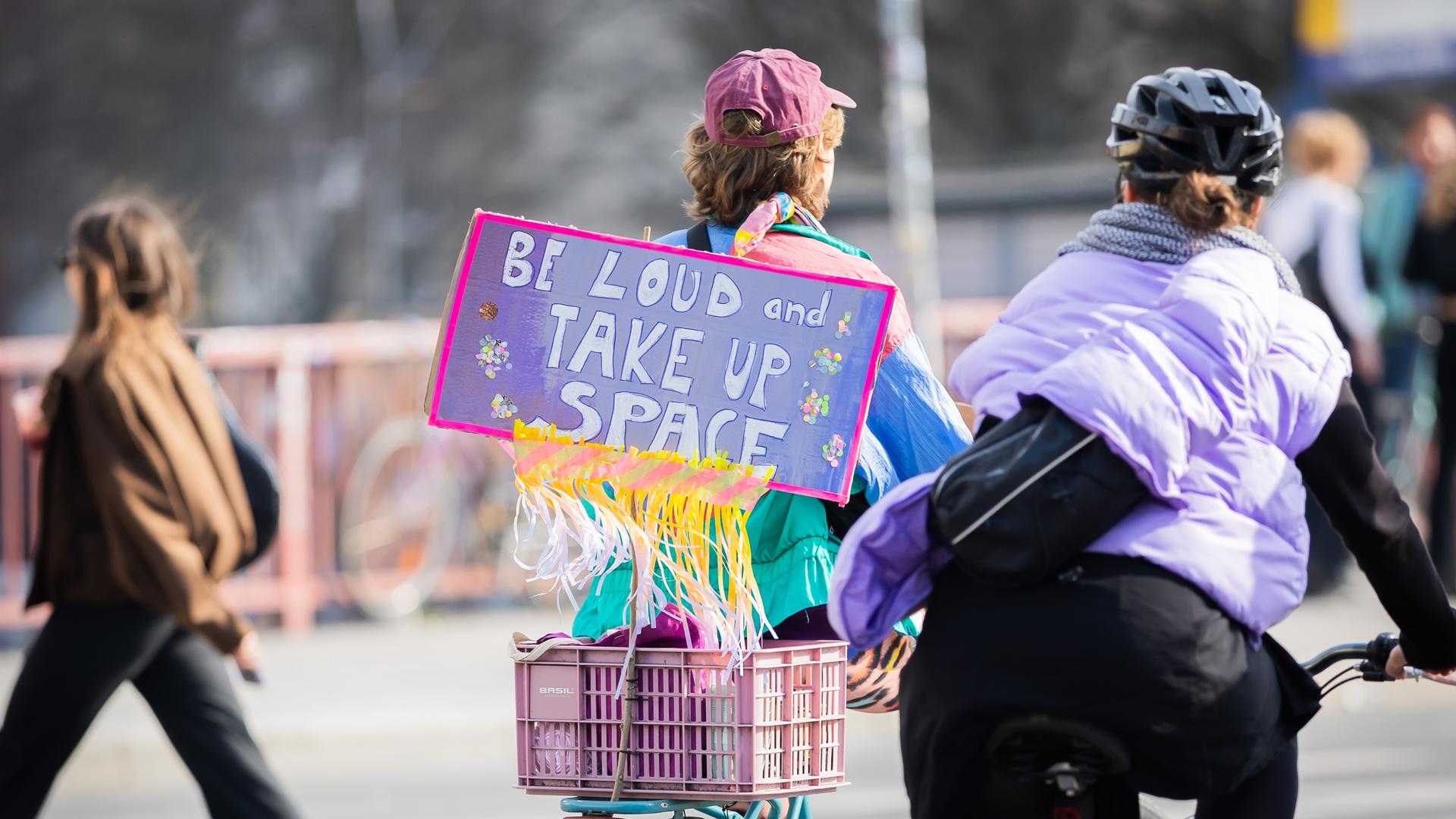 Zwei Teilnehmerinnen einer Frauen-Fahrrad-Demo in Berlin. Auf ihrem Schild steht "Be loud and take up space" (Sei laut und nimm Raum ein). 