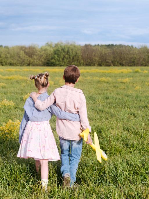 Ein Junge und ein Mädchen halten sich an den Händen und laufen über ein Feld. Der Junge hält ein Spielzeug-Flugzeug in der Hand. Man sieht die Kinder von hinten. 