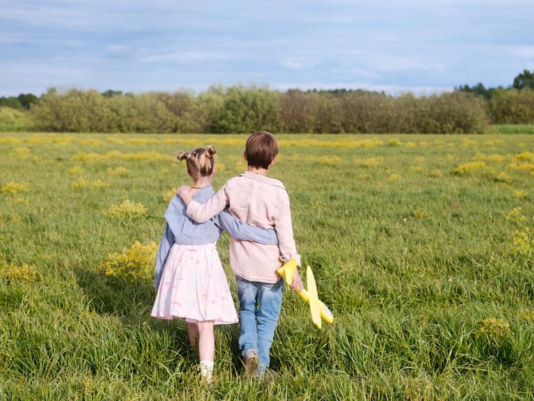 Ein Junge und ein Mädchen halten sich an den Händen und laufen über ein Feld. Der Junge hält ein Spielzeug-Flugzeug in der Hand. Man sieht die Kinder von hinten. 