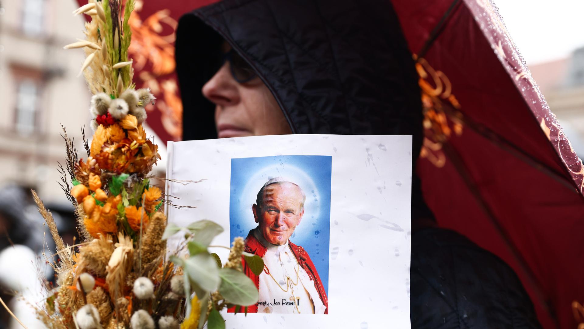 Eine Person mit Sonnenbrille und schwarzer Kapuze trägt eine Illustration von Papst Johannes Paul II. und einen Strauss mit Trockenblumen während einer Demonstration in Krakow.