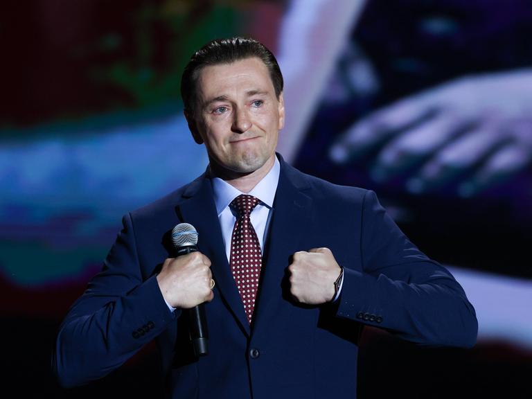 Sergei Besrukow steht mit dem Mikrofon in der Hand auf einer Bühne und macht einen bewegten Gesichtsausdruck.