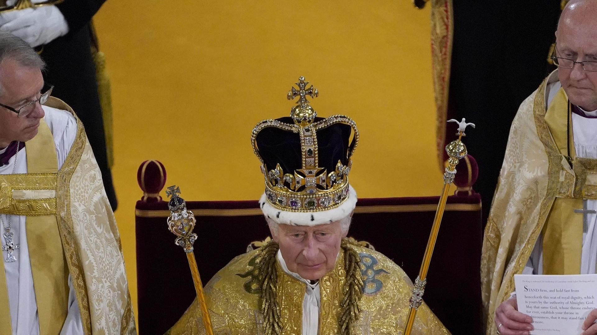 König Charles III. sitzt in der Westminster Abbey auf einem Stuhl, auf dem Kopf trägt er die Krone, in den Händen hält er das Zepter und den Stab.