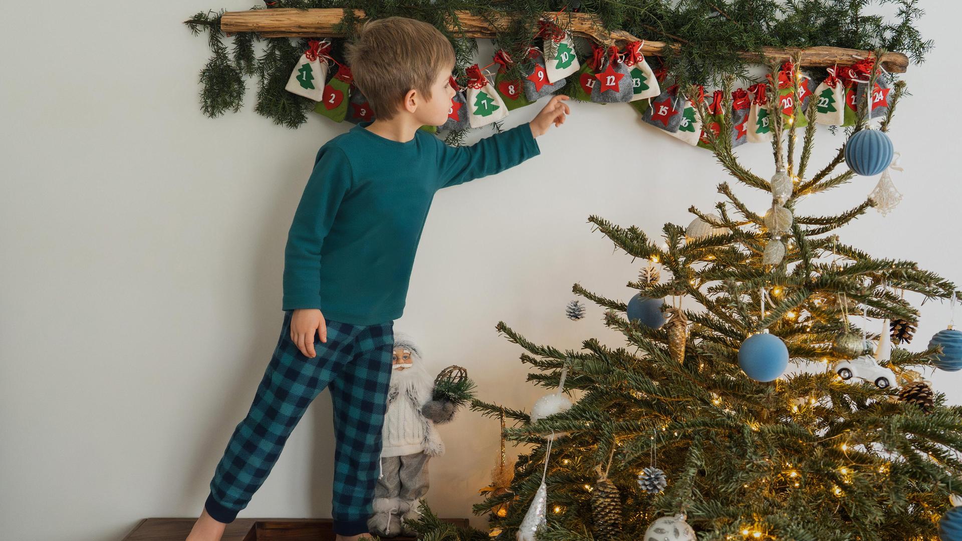 Ein Junge greift nach einem Säckchen im Adventskalender, neben ihm steht ein kleiner geschmückter Weihnachtsbaum