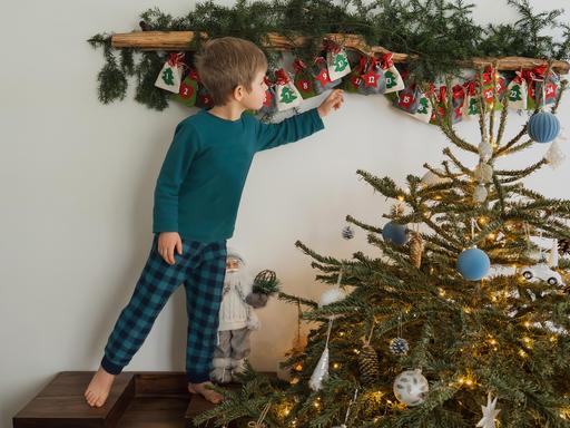 Ein Junge greift nach einem Säckchen im Adventskalender, neben ihm steht ein kleiner geschmückter Weihnachtsbaum