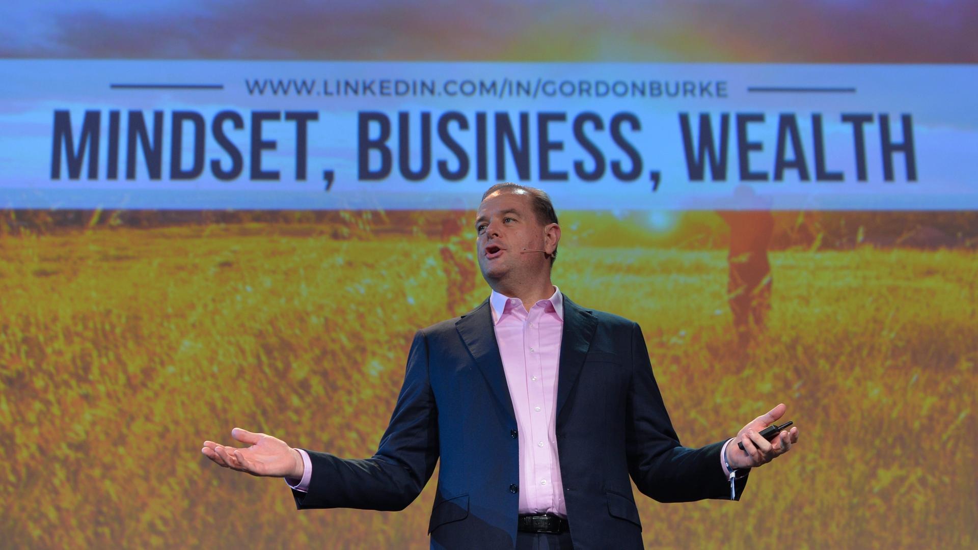 Ein Mann im Anzug steht mit ausgebreiteten Armen auf einer Bühne. Hinter ihm wird das Bild einer sonnenbestrahlten Wiese projiziert, darauf die Worte: "Mindset, Business, Wealth".