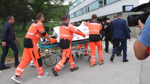 Mehrere Rettungssanitäter in organgefarbener Kleidung schieben die Bahre mit Fico zum Krankenhaus. Sie halten ein weißes Tuch hoch, damit Fico nicht zu sehen ist. 
