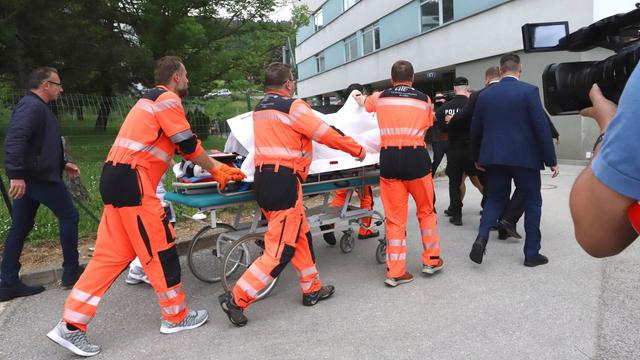 Mehrere Rettungssanitäter in organgefarbener Kleidung schieben die Trage mit Robert Fico zum Krankenhaus. Sie halten ein weißes Tuch hoch, damit Fico nicht zu sehen ist. 