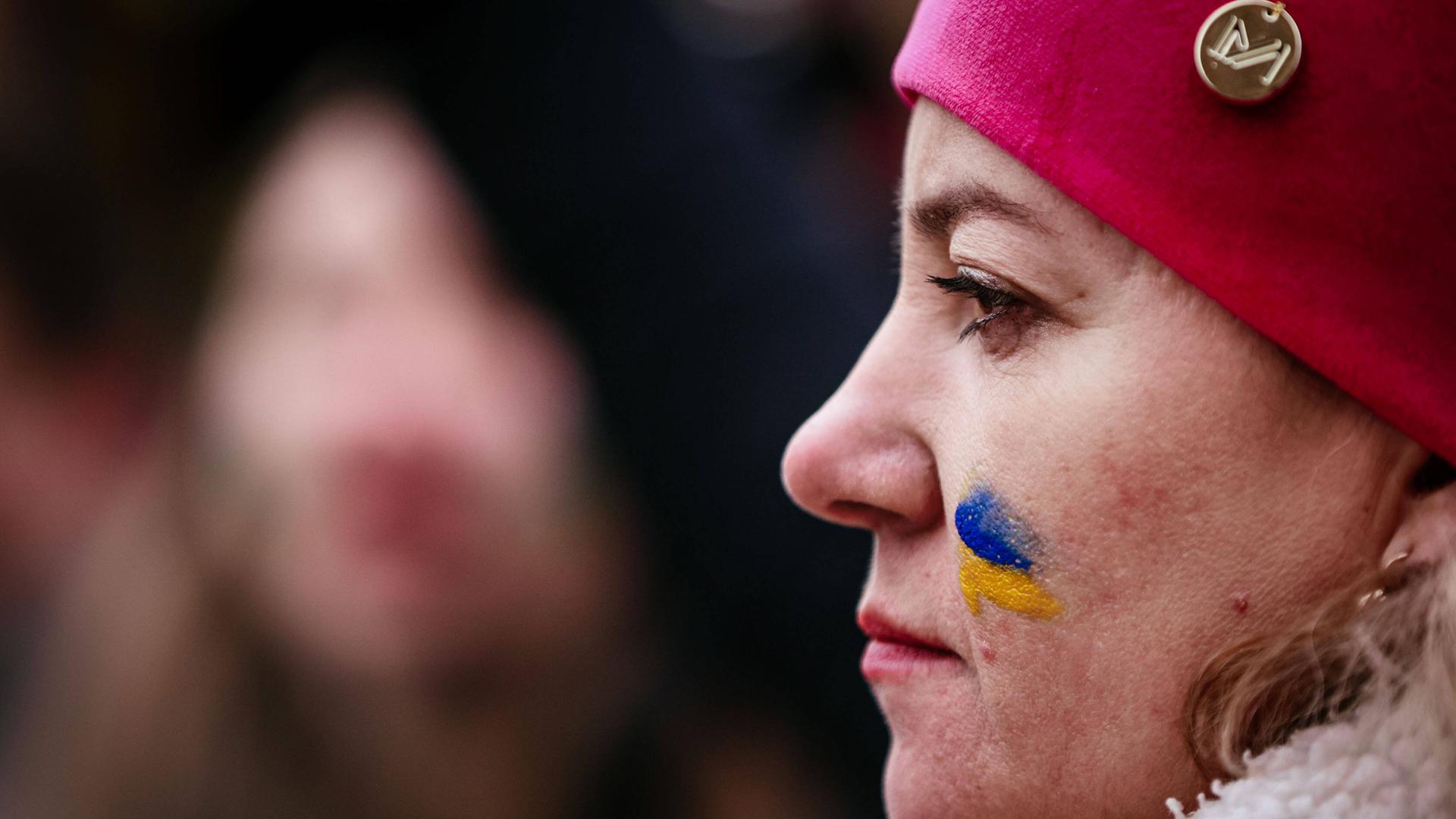 Profil einer Demonstrantin mit einer roten Mütze und einer gemalten ukrainischen Flagge auf der Wange.